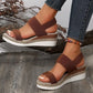 Brown espadrille wedge sandals with elastic straps and a gold-tone ankle chain, shown on a model in a lifestyle setting with natural elements.