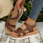 Elegant brown faux leather women's flat sandals with knot detail and adjustable hook-and-loop straps, featuring a wood-grain platform sole, shown worn on a foot in a natural outdoor setting.
