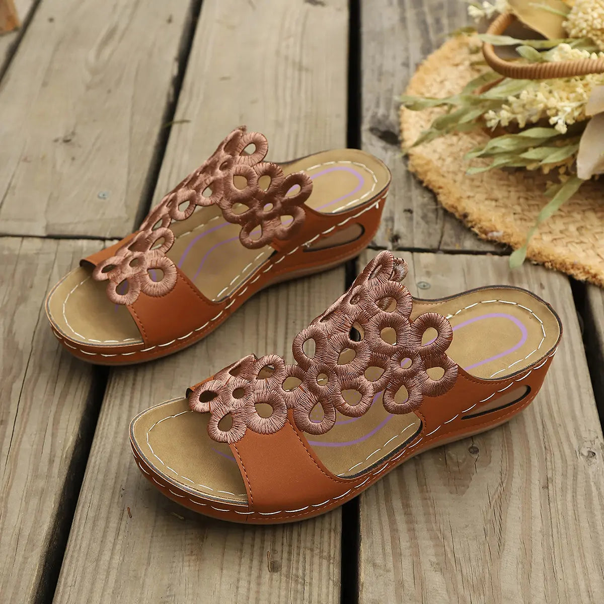 Brown faux leather wedge sandals for women featuring intricate laser-cut floral patterns on the wide strap, with a comfortable cushioned footbed and wedge heel, styled on a rustic wooden surface with a straw hat accessory.