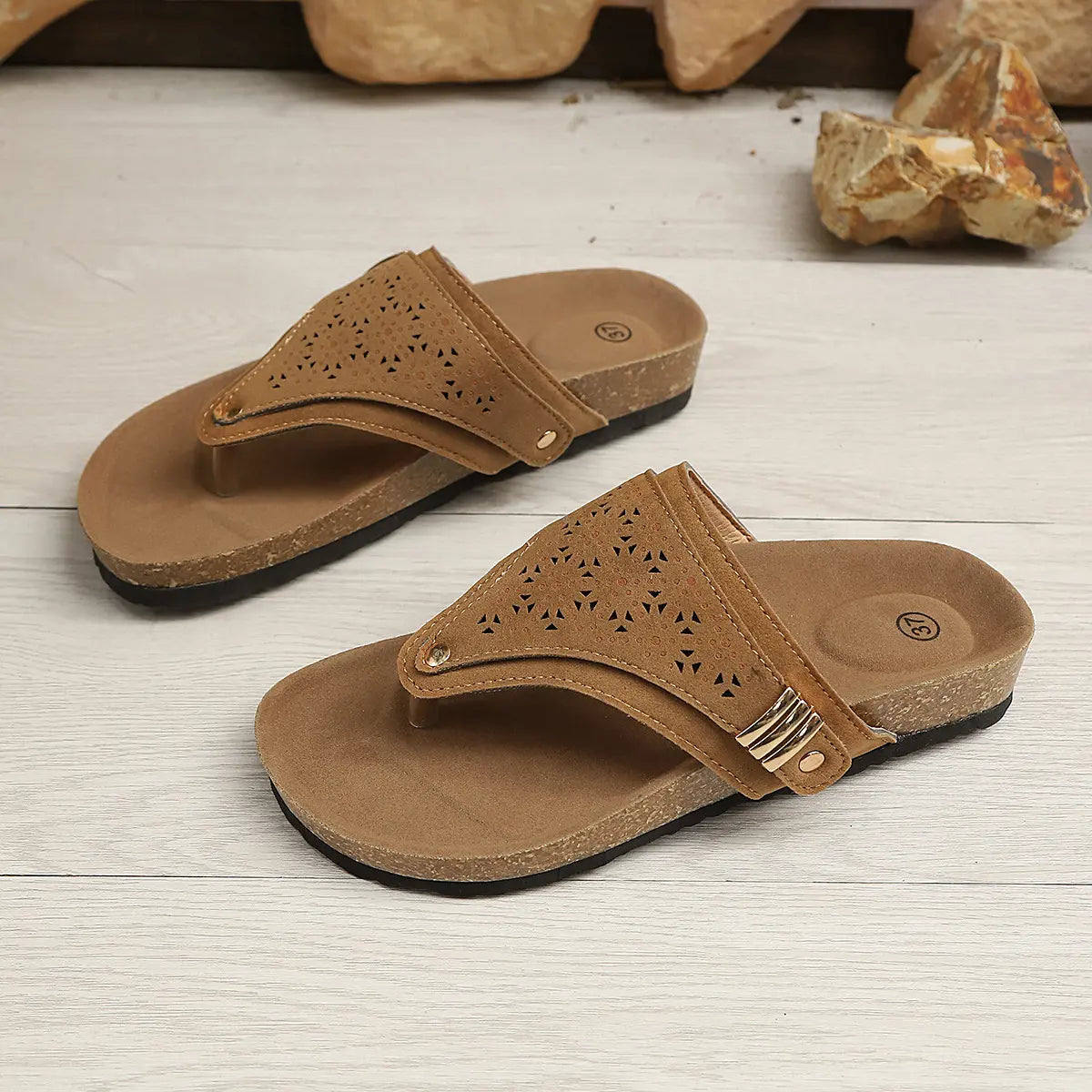 Brown suede-like thong sandals with intricate laser-cut patterns, cork-textured footbeds, and gold buckle accents, shown from a three-quarter angle on a wooden surface with decorative rocks.