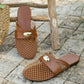 Brown mesh pointed-toe flat mules with a dark brown faux leather strap and polished gold oval hardware, resting on a stone surface with a straw bag accessory.
