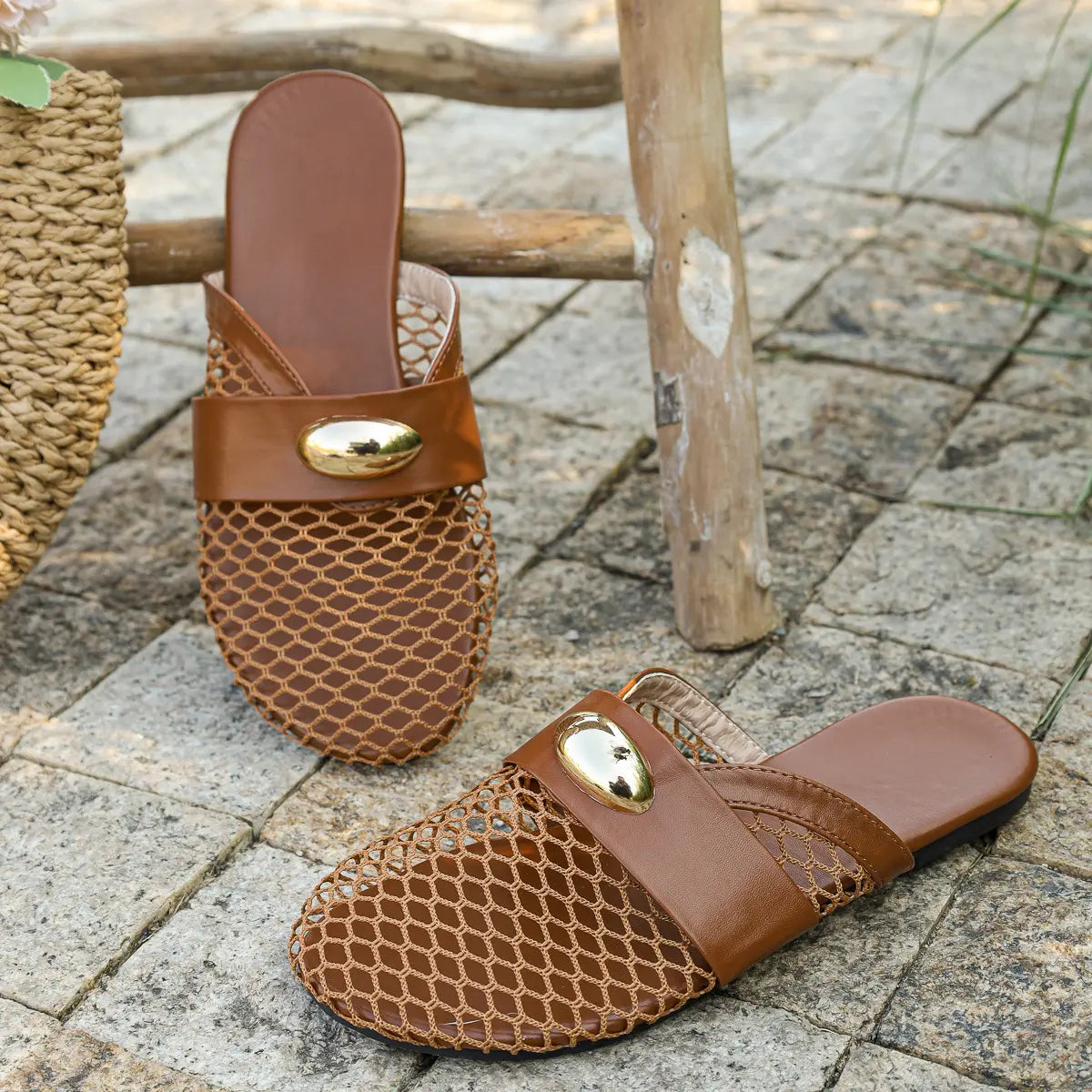 Brown mesh pointed-toe flat mules with a dark brown faux leather strap and polished gold oval hardware, resting on a stone surface with a straw bag accessory.
