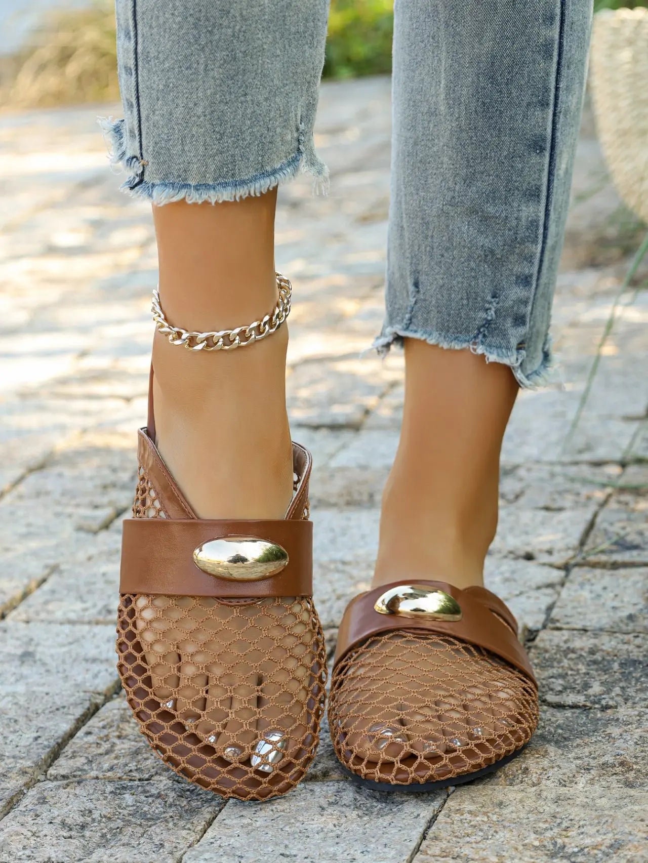 Women's brown mesh pointed-toe flat mules with a faux leather strap and polished gold oval hardware, styled with distressed denim jeans on a paved outdoor surface.