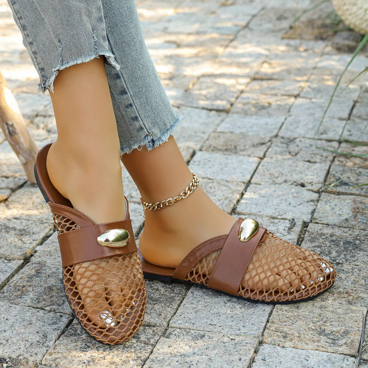 Brown mesh pointed-toe flat mules worn by a model with distressed denim jeans and a gold anklet, showcasing the intricate diamond weave and gold oval hardware detail on a stone patio.