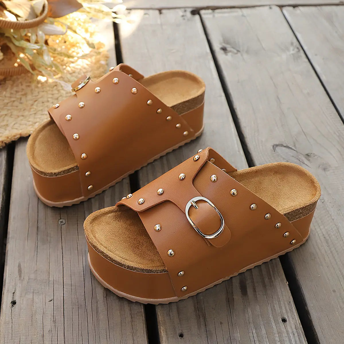 Brown studded platform slide sandals with gold buckle and cork-effect footbed, shown from a three-quarter angle on a wooden deck.