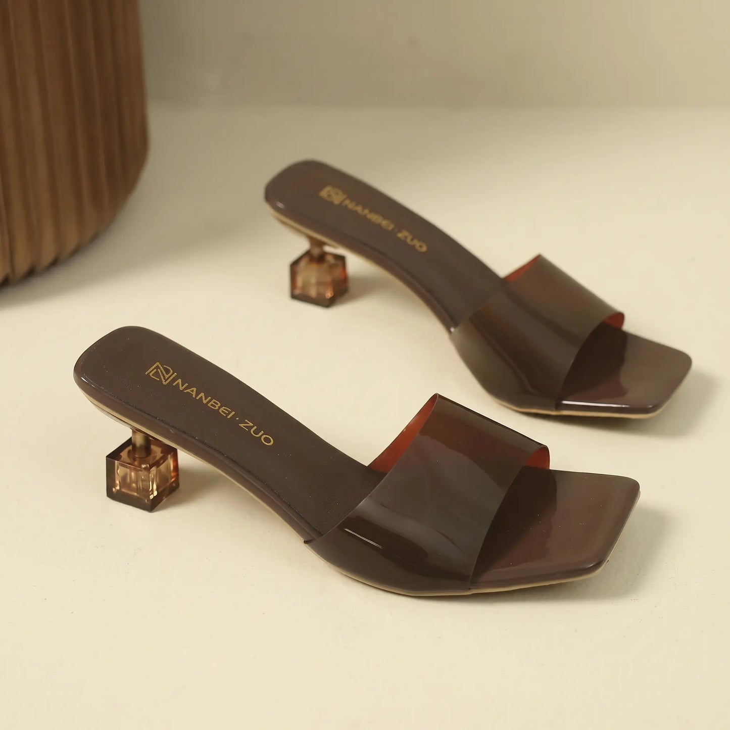 A pair of dark brown transparent slip-on mule sandals with square toes and geometric block heels, showcasing the NANBEI ZUO brand detail on the footbed.