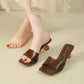 Close-up of a woman's feet wearing brown transparent strap square toe slip-on mule sandals with clear geometric block heels. One sandal is worn, the other rests beside it on a light beige surface.