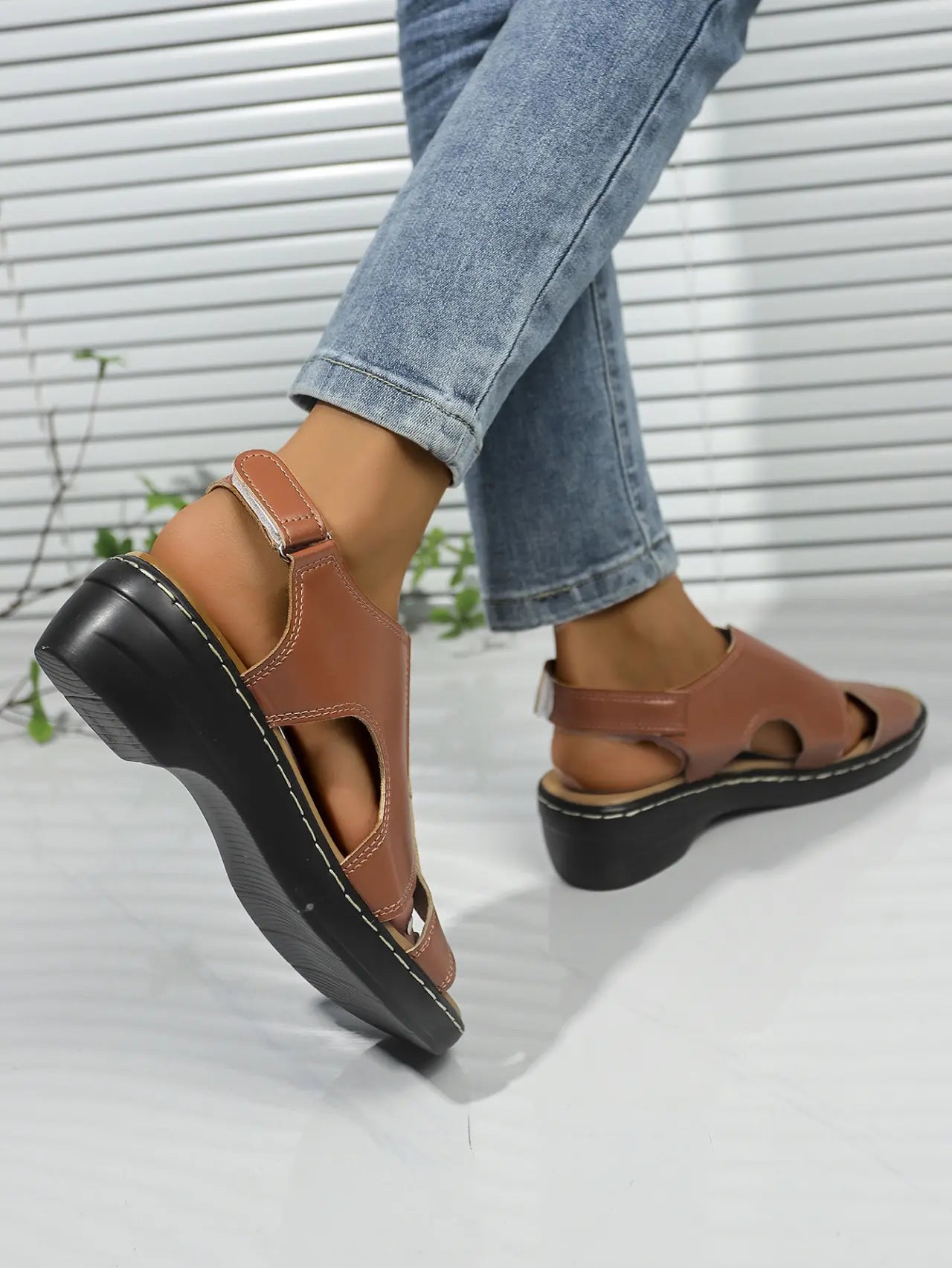 Model wearing elegant brown faux leather peep-toe wedge sandals with adjustable ankle strap and white contrast stitching, paired with denim jeans.