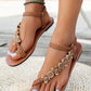 Bohemian brown woven flat sandals with decorative wood-effect floral and circular embellishments on the T-strap, worn on feet with white toenails and a turquoise beaded anklet.