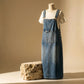 Front view of Brynox medium blue denim midi pinafore dress on a mannequin, showcasing adjustable straps, multi-compartment bib pocket, and side buttons. Features a faded wash and contrast stitching.