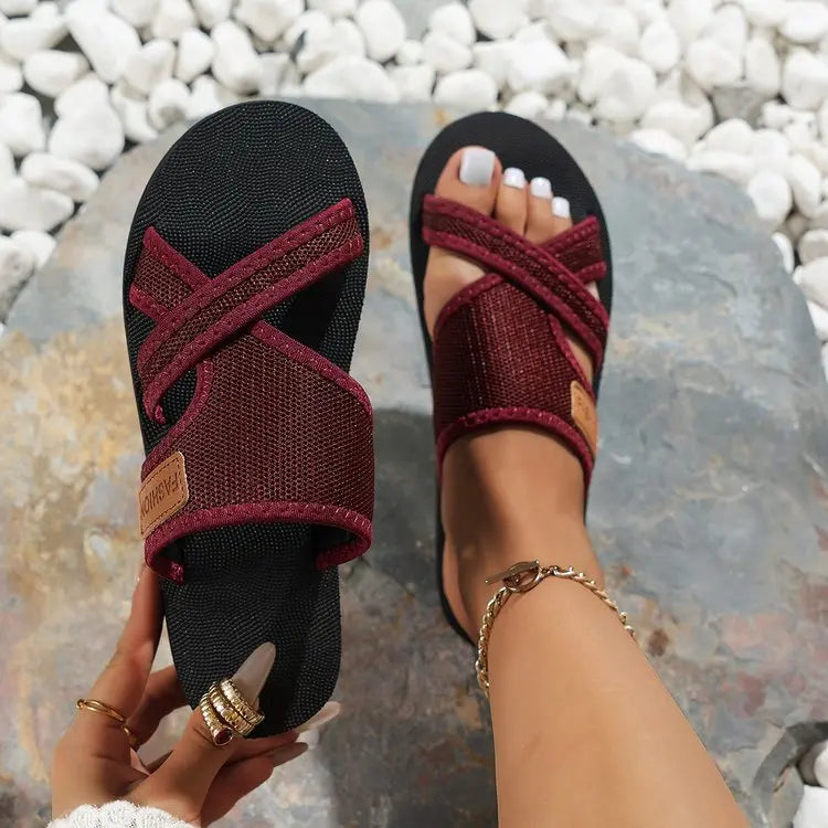Women's burgundy fabric criss-cross platform slide sandals worn on feet, showcasing contoured footbed and textured upper with a small 'FASHION' label. Set on natural pebbles and stones for a chic summer look.