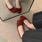 Top-down view of elegant burgundy faux suede ballet flats worn on feet, featuring classic bow details and square-round toes. Reflected in a mirror on a textured rug.