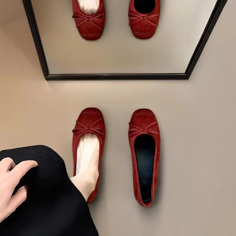 Burgundy faux suede ballet flats with a delicate bow and square-round toe, one worn on a foot and reflected in a mirror.