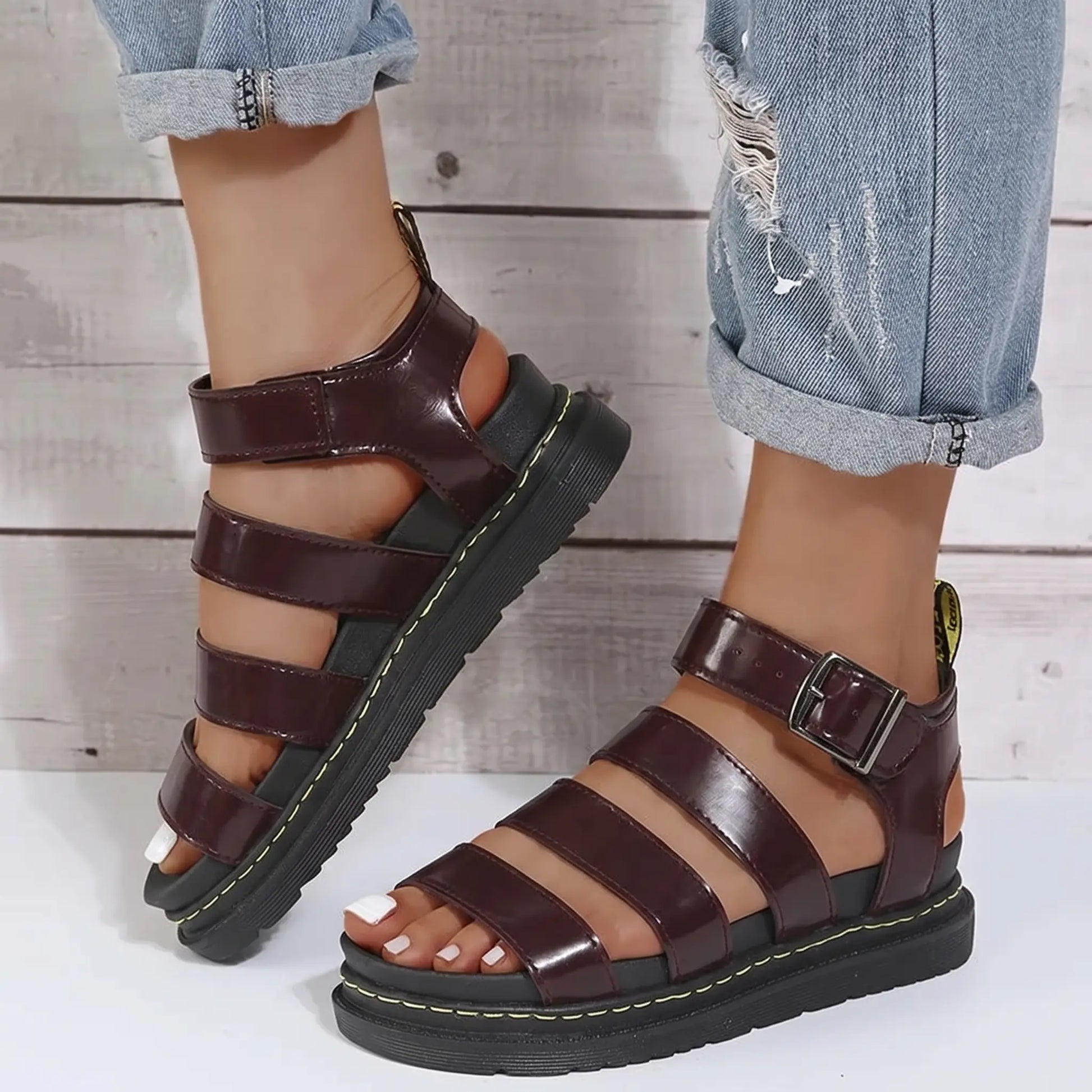 A pair of deep burgundy PU leather gladiator sandals with a chunky black platform sole and yellow stitching, worn by a person in cuffed denim jeans.