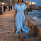 Woman wearing a Zivro chambray blue A-line midi shirt dress with cuffed sleeves and a gold buckle belt, walking by the sea.