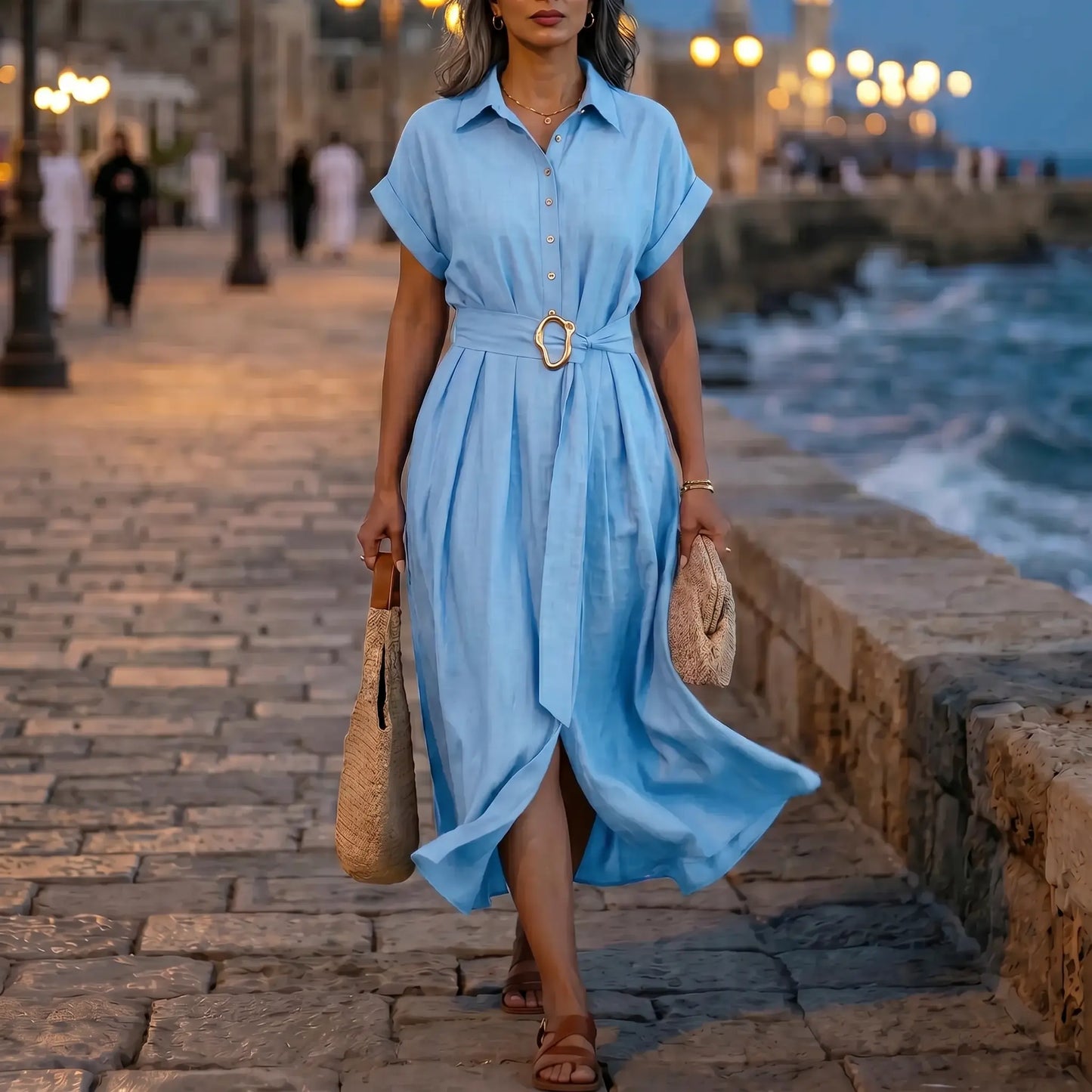 Woman wearing a Zivro chambray blue A-line midi shirt dress with cuffed sleeves and a gold buckle belt, walking by the sea.