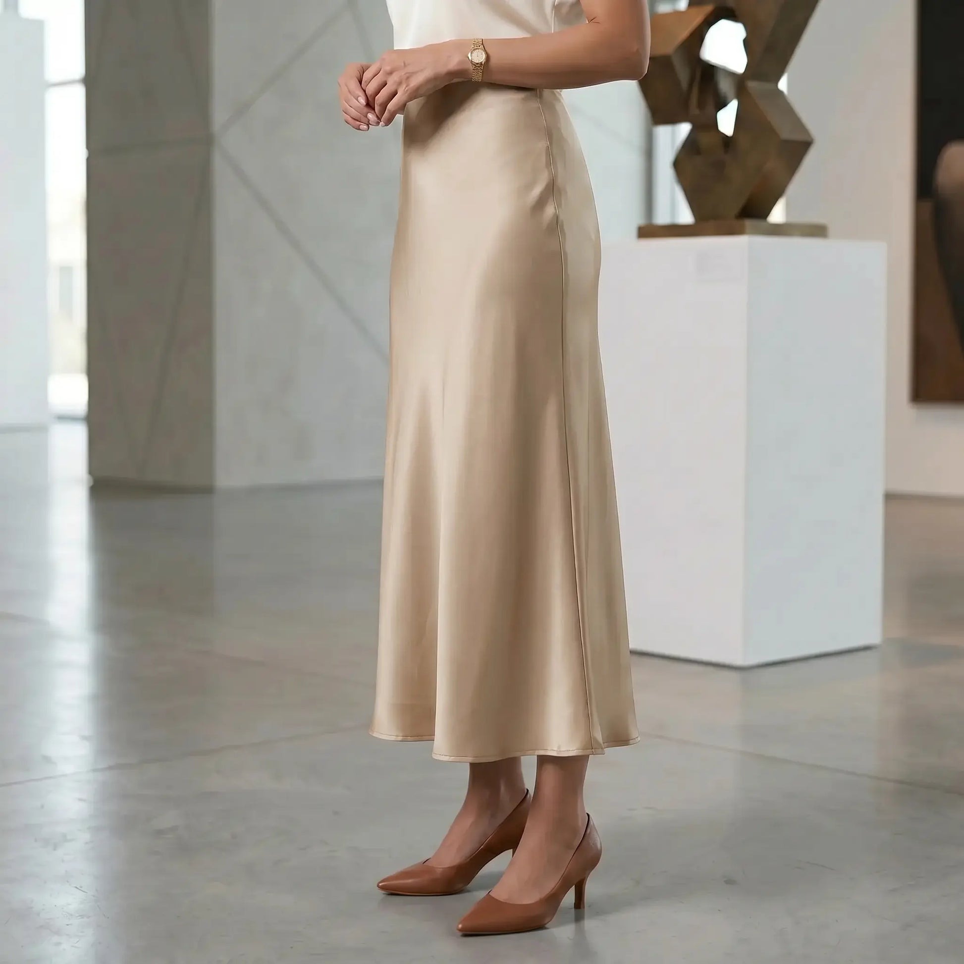 Woman wearing a high-waisted champagne beige satin midi skirt with a fluid A-line mermaid drape, styled with brown heels, in a modern art gallery setting.