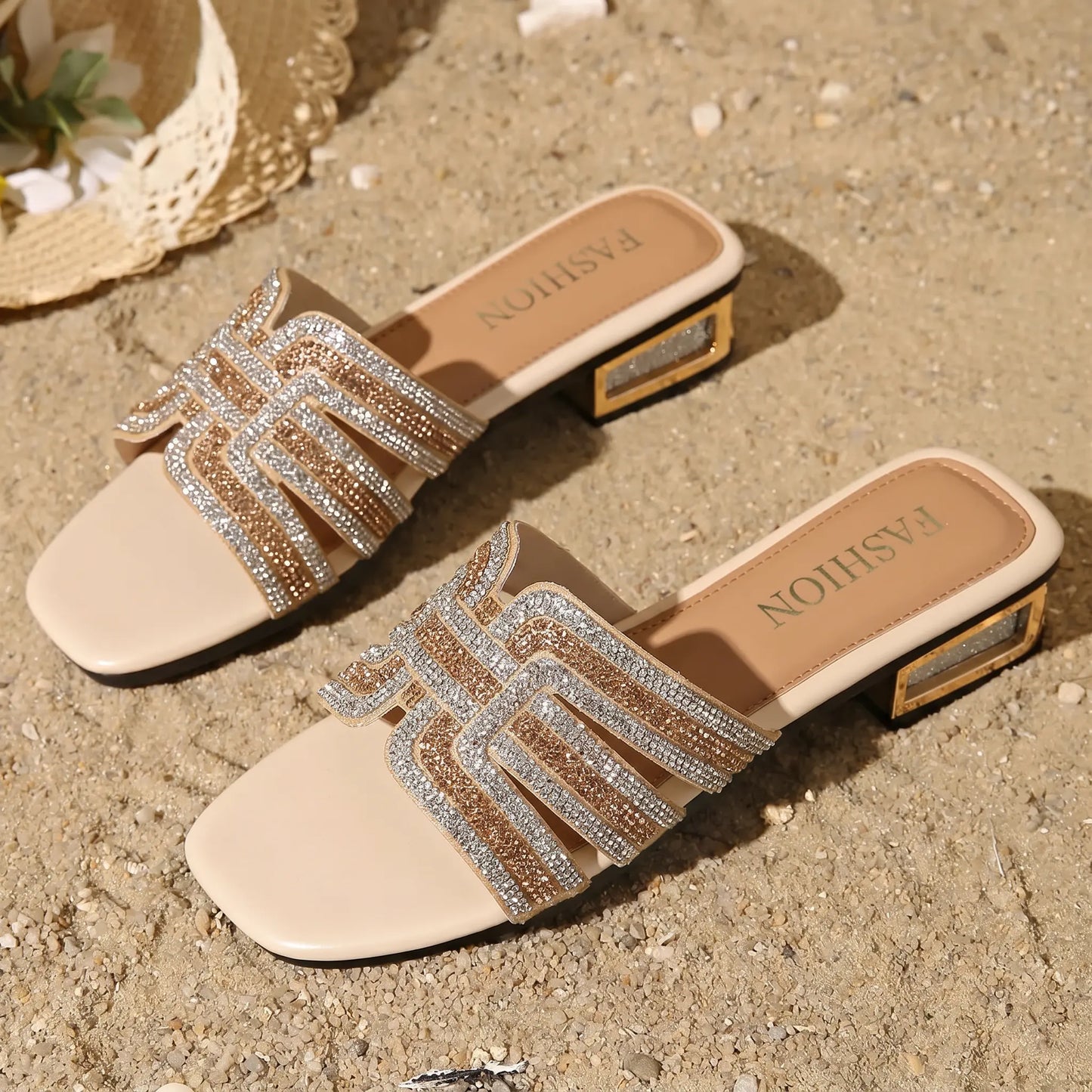 Champagne shimmer embellished flat slide sandals for women, featuring a square toe, intricate geometric rhinestone straps, and a low block heel with metallic glitter accents, displayed on a sandy beach with a straw hat.