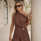 A woman models a chic chocolate brown linen-blend two-piece set featuring an asymmetrical one-shoulder top with a draped bodice and gold buckle accent, paired with high-waisted shorts, styled outdoors.