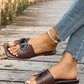 Chic chocolate brown square-toe flat slide sandals adorned with a decorative initial buckle featuring crystal accents, shown on a woman's foot with distressed denim.