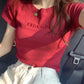 Female model wearing a Classic Crimson slim-fit stretch jersey t-shirt with tonal embroidered 'MISCHIEF' text, paired with light trousers and a gold necklace, seated in a car.