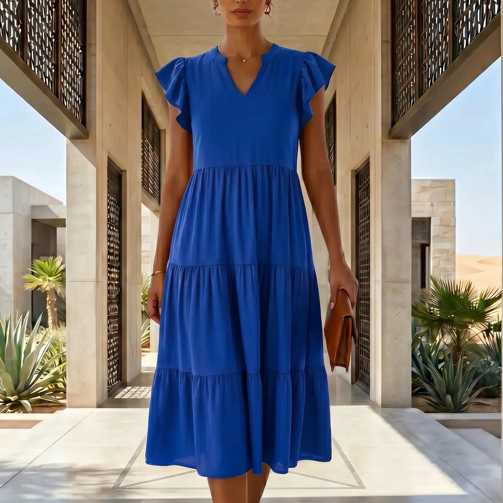 Model wearing a Torvun Cobalt Blue Ruffled Tiered Midi Dress with V-neck and flutter cap sleeves, walking in a modern architectural desert setting.