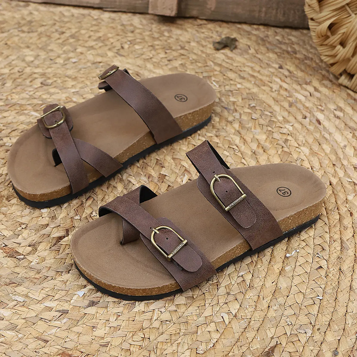 Brown cork footbed slide sandals with dual matte suede-effect straps and antique brass buckles, presented on a woven straw mat.