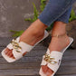 Cream flat slide sandals featuring a chunky gold chain across the vamp and a modern square toe, styled on a person's feet with denim jeans in an outdoor setting.
