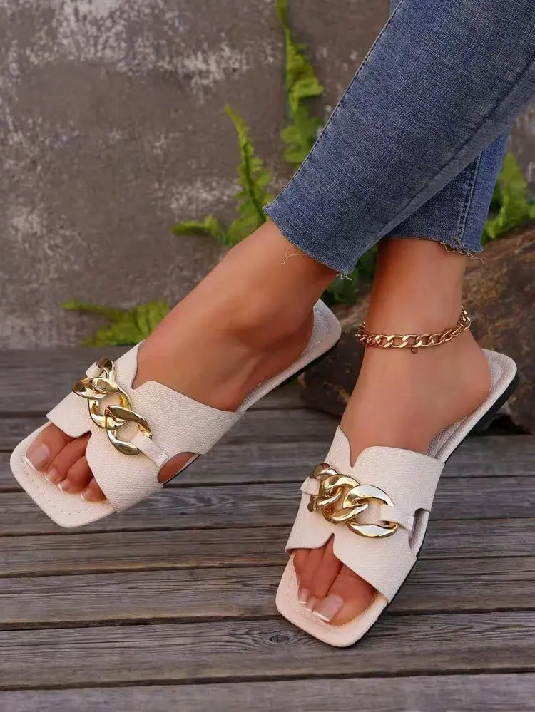 Cream flat slide sandals featuring a chunky gold chain across the vamp and a modern square toe, styled on a person's feet with denim jeans in an outdoor setting.