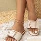 Cream flat slide sandals on feet featuring a wide strap with a chic gold-tone square buckle and a modern square toe, set against a textured lifestyle background.