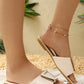 Elegant cream flat slide sandals adorned with a chic square gold-tone buckle, shown on a model's feet in a relaxed outdoor setting with decorative plants.