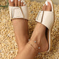 Elegant cream flat slide sandals with a chic square gold-tone buckle, shown on feet with white toenail polish, styled on a textured pebble background.