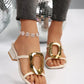 Women's cream ivory slide sandals with a large gold oval buckle and a comfortable block heel, worn on a model's feet with sparkly grey pants. Sandals feature a square toe and thin toe post strap.
