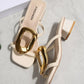 Creamy ivory square-toe slide sandals featuring a large, polished gold oval buckle and a comfortable block heel, styled on a textured studio background.