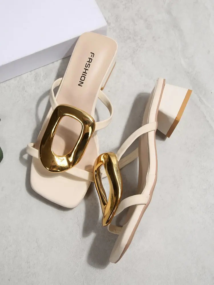Creamy ivory square-toe slide sandals featuring a large, polished gold oval buckle and a comfortable block heel, styled on a textured studio background.