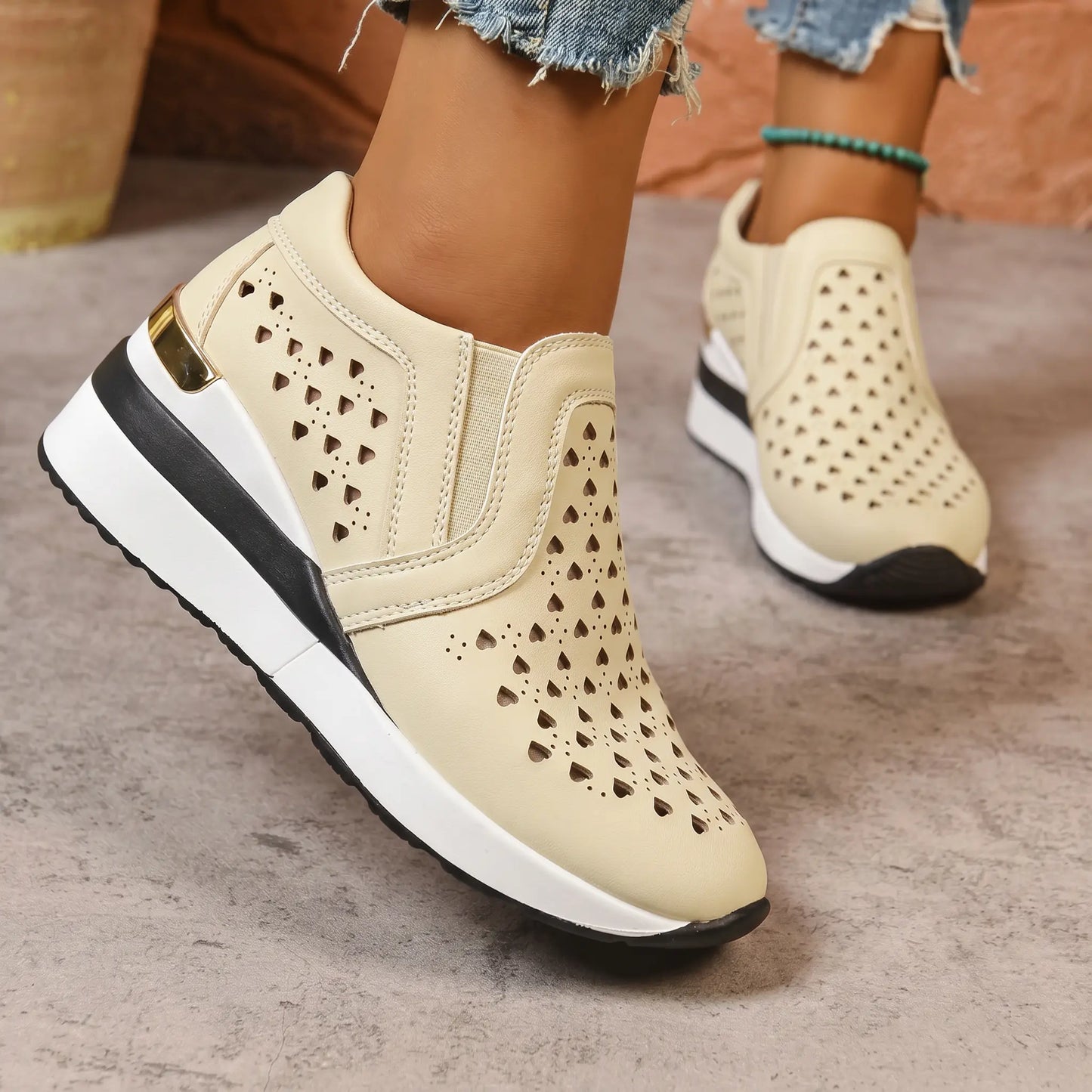 Warm cream perforated wedge sneakers worn with ripped jeans, showcasing the heart pattern, gold heel accent, and stacked sole from a three-quarter view.