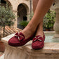 Women's crimson red faux-suede moccasin loafers featuring a prominent gold metallic bow, worn elegantly by a person in an outdoor courtyard setting near a fountain.
