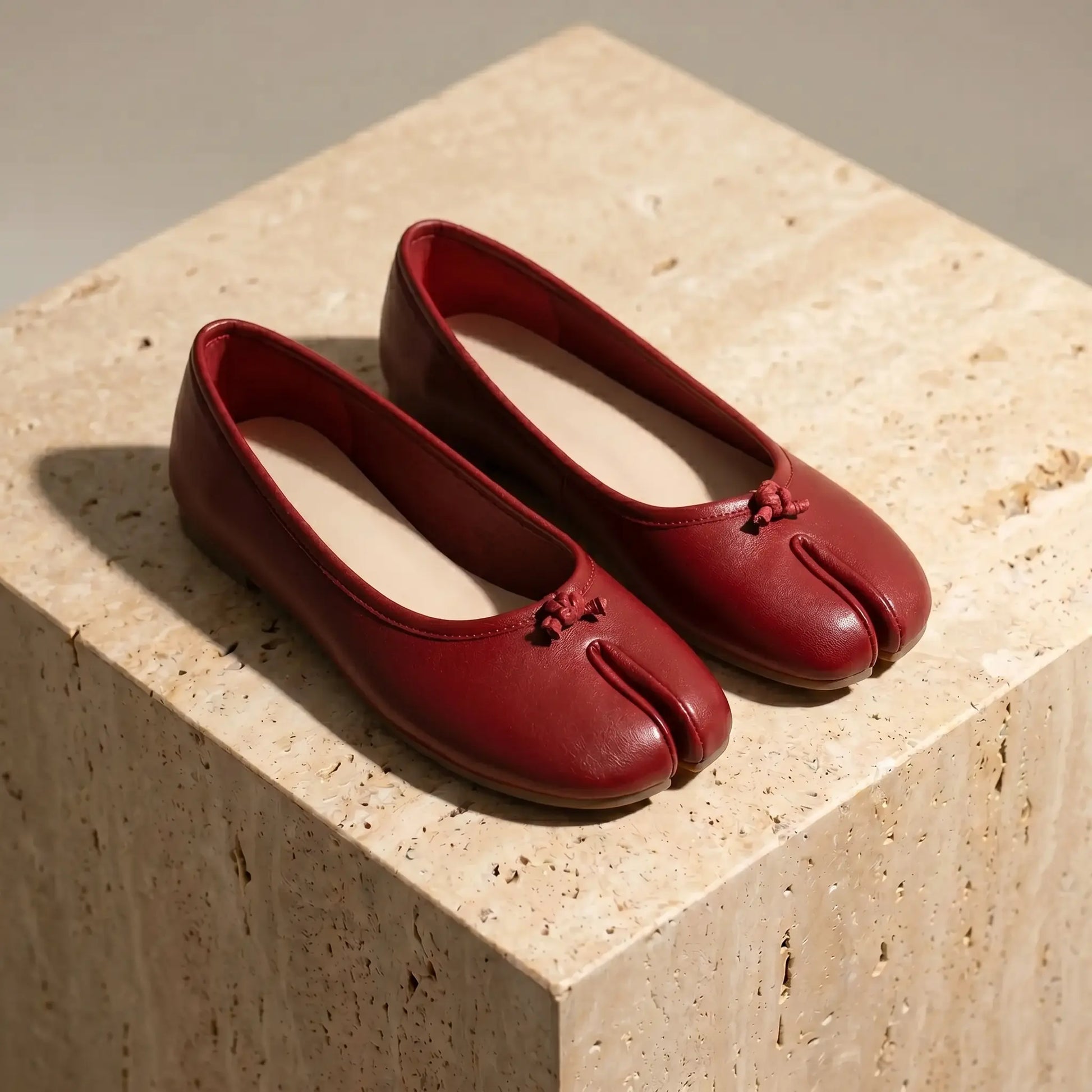 Pair of crimson red vegan leather split-toe ballet flats with delicate bows, displayed from a three-quarter angle on a textured beige stone block.
