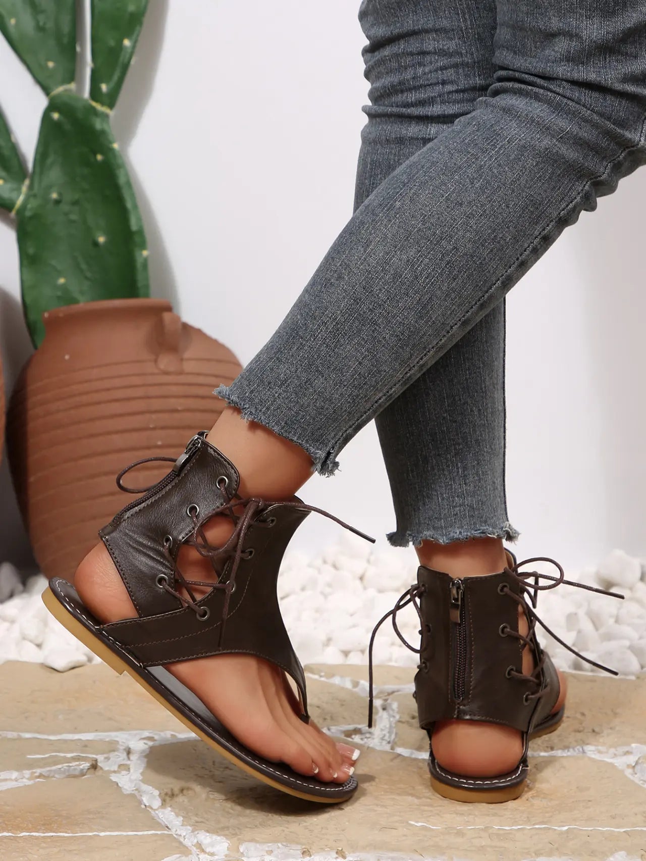 Dark brown faux leather gladiator flat sandals for women, featuring intricate lace-up detailing extending up the ankle and a thong toe post, worn with denim jeans.