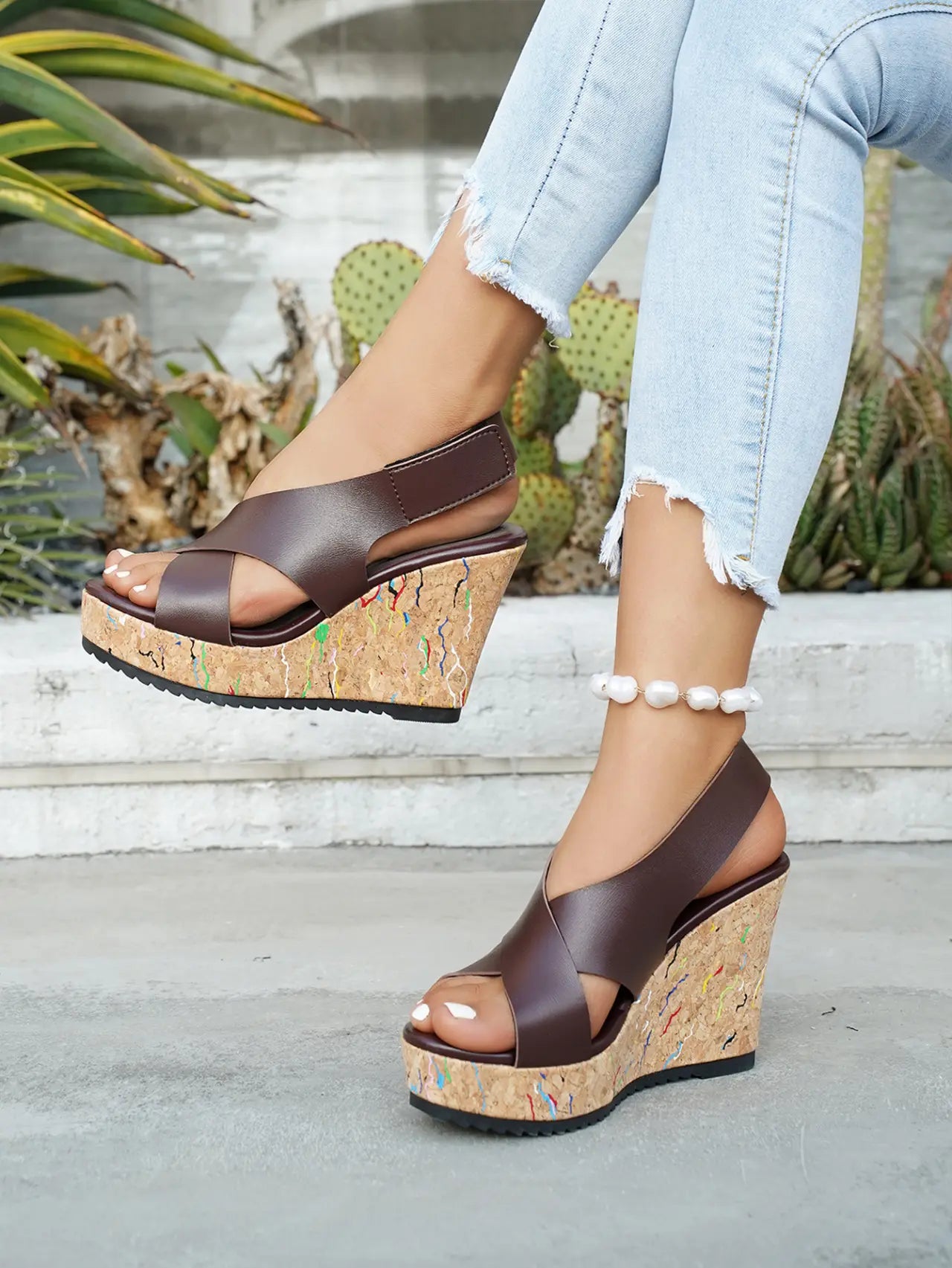 Women's dark brown criss-cross strap wedge sandals with a multi-color speckled cork platform and wedge, shown worn on feet with light wash denim jeans.