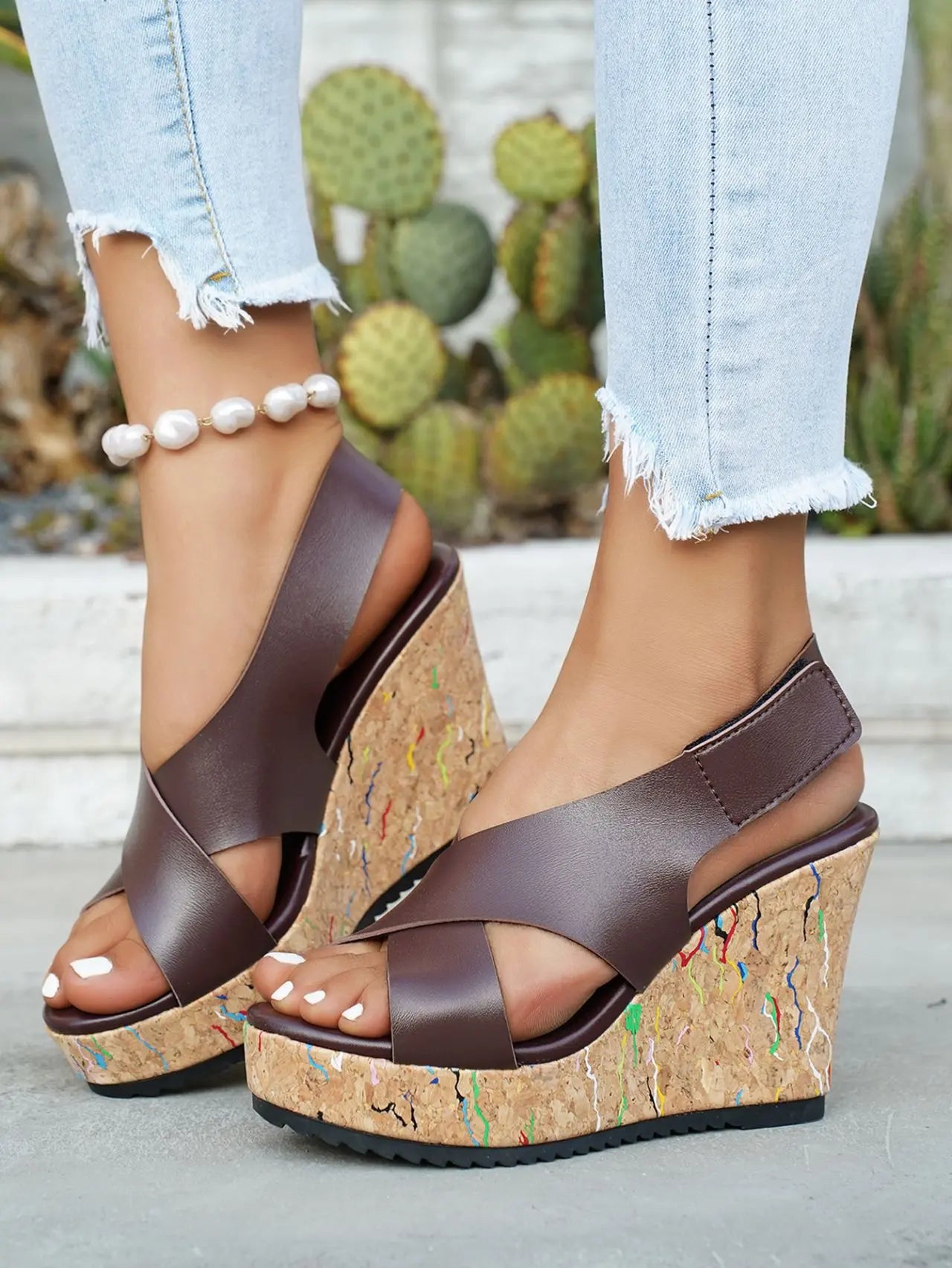 Model's feet wearing dark brown faux leather criss-cross wedge sandals with vibrant multi-color speckles on the cork wedge, styled with light wash jeans.