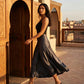 Woman elegantly walking in a Deep Onyx high-waisted satin midi skirt, showcasing its fluid A-line mermaid drape against an ornate architectural background at golden hour.