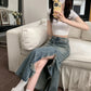 Model sitting on a white sofa, showcasing the Zendru Asymmetrical Ruffled Denim Midi Skirt. This vintage blue, high-waisted skirt features a dramatic cascading ruffled slit that reveals the leg.