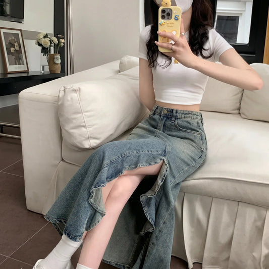 Model sitting on a white sofa, showcasing the Zendru Asymmetrical Ruffled Denim Midi Skirt. This vintage blue, high-waisted skirt features a dramatic cascading ruffled slit that reveals the leg.