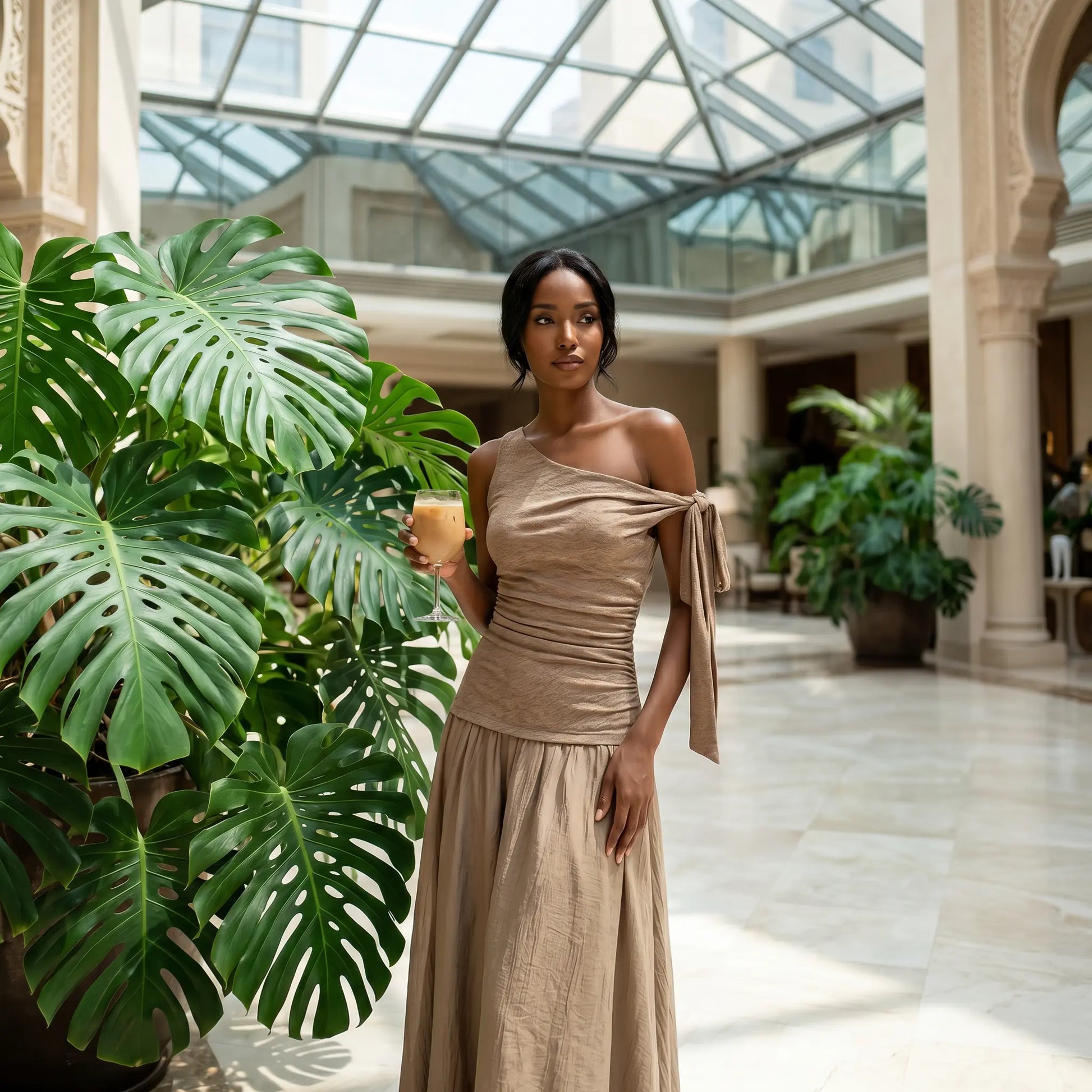 Elegant model in a Zolwen coffee taupe asymmetric one-shoulder ruched drop-waist maxi dress, posing gracefully in a sunlit atrium.