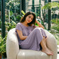 Woman relaxing in a dusty lavender Zevryn romantic lounge dress, featuring a gathered scoop neck with a delicate tie and subtle heart pointelle pattern, in a sunlit indoor garden.