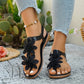 Black Dyraen bohemian floral flat sandals for women, featuring layered fabric flower embellishments on the vamp and an elasticated slingback strap, worn with light denim jeans in an outdoor setting.