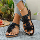 Black flat thong sandals adorned with layered fabric floral appliqués and a comfortable elasticated slingback strap, worn on feet with white nail polish in an outdoor setting.