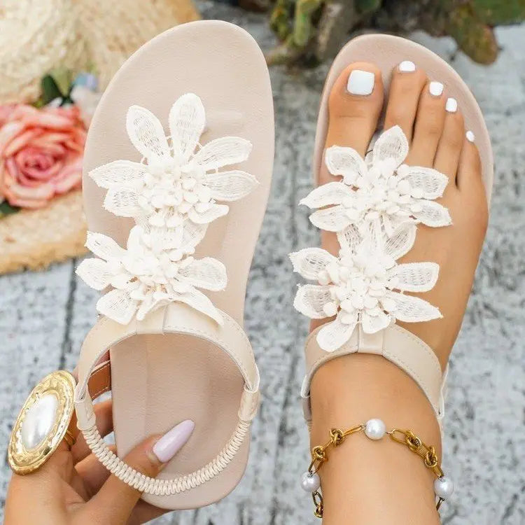 Women's flat beige thong sandals adorned with layered white fabric floral appliques, featuring an elasticated slingback strap, shown on a model's feet with white toenails and a pearl bracelet.