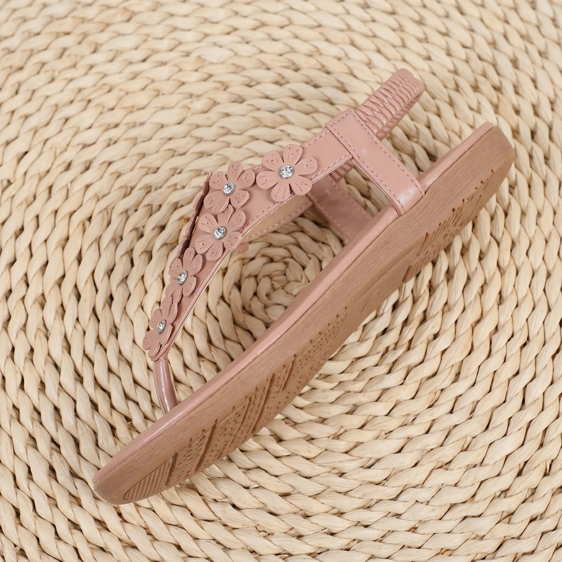 Blush apricot Elaraia t-strap flat sandals with 3D floral appliques and clear rhinestone accents, featuring an elasticated slingback strap and a textured rubber sole, presented on a woven mat.